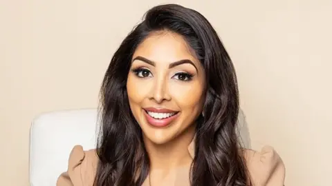 Dr Vanita Rattan Dr Rattan smiling at the camera, wearing a light brown blazer, sitting on a white chair. The background is plain light brown.