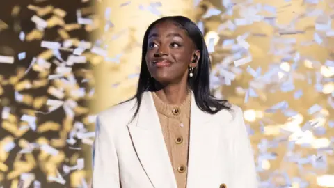 Getty Images Cat on stage, she's wearing a white suit over a beige jumper, the stage backdrop is gold and confetti falls around her.