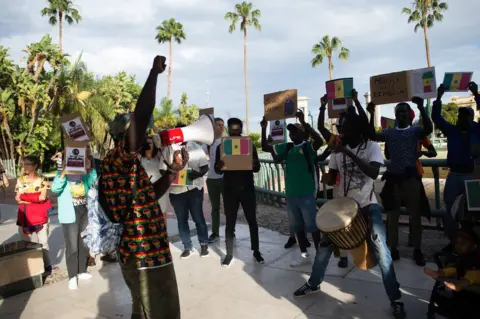 Jesus Merida/ Getty Images Senegalese protesters holding placards outside next to tall palm trees. One man is talking through a speakerphone and the other men appear animated - 7 June.