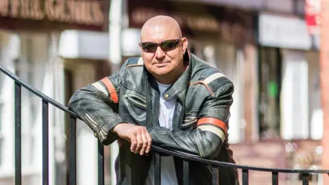 Phil Rigby Mike Craven wearing sunglasses and a leather jacket leans on a metal fence