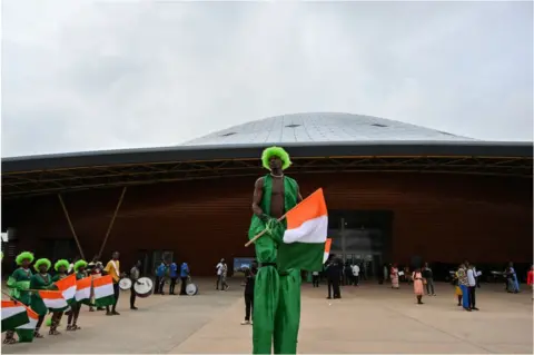 Issouf Sanogo/ AFP Man in stilts wearing Ivorian flag colours and a green wig.