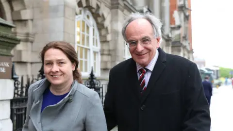 ISABEL INFANTES/GETTY IMAGES Woman with dark hair wearing grey coat and man with grey hair and glasses wearing dark coat and tie