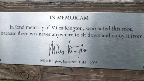 OpenBenches Bench dedication: "In fond memory of Miles Kington, who hated this spot, because there was never anywhere to sit down and enjoy it from."