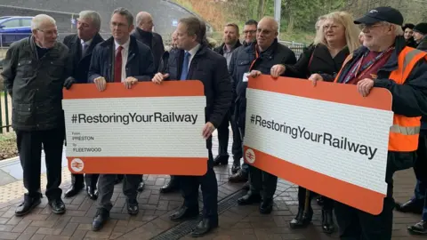 BBC Grant Shapps and people holding giant rail tickets on the platform at Fleetwood