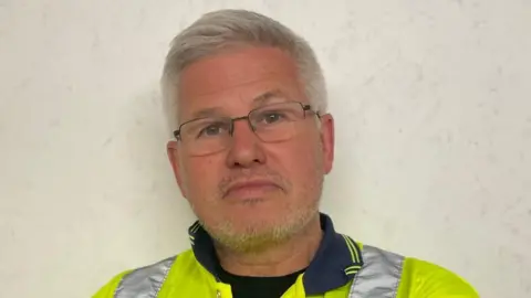 Andrew McCausland Landlord Andrew McCausland stands in a high vis jacket arms folded. He has silver cropped hair and light stubble. He's wearing glasses and looking at the camera.