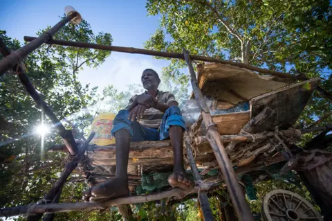 Roshni Lodhia A portrait showing a fisherman sitting on a stilted structure in a mangrove forest