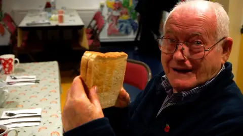 An elderly man smiles as he holds a sandwich