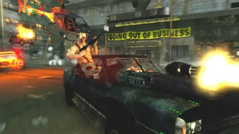 Sony Computer Entertainment A chaotic screenshot from a video game shows a shirtless man in a clown mask sitting on the lip of a car door as he holds a large shotgun. The modified sports car he's a passenger in is fitted with two large gatling gun weapons - a bright yellow muzzle flash emanates from one. Behind him is a post-apocalyptic scene - we can see a building with a large, yellow "going out of business" banner across its front. A helicopter flies through the shot, a parked car appears to be on fire and a third, modified vehicle appears to be chasing the car carrying the man with the shotgun.