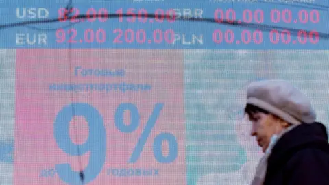 Getty Images EBRUARY 24, 2022: A woman walks past a digital board displaying the current rates outside a currency exchange office. As of 10am, the Moscow Exchange has reported the US dollar and Euro trading at 89.6 and 99.99 respectively against the Russian rouble, thus reaching their historic highs. Vitaly Nevar/TASS