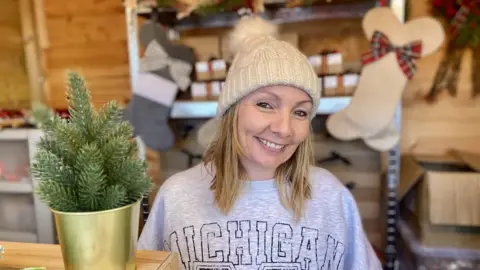 BBC Woman wearing bobble hat smiling at camera from inside a wooden stall