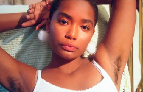 Billie on Unsplash A young black woman poses with her arms above her head, showing her armpit hair