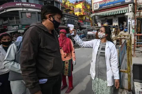 AFP A volunteer (R) checks the temperature of devotees who arrive to take a holy dip in the waters of the River Ganges during Makar Sankranti, a day considered to be of great religious significance in the Hindu mythology, on the first day of the religious Kumbh Mela festival in Haridwar on 14 January 2021