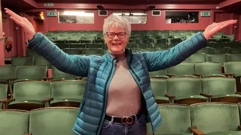 BBC Anne Lane is stood inside the auditorium at the Norbury Theatre in Droitwich - behind her are rows of green seats.