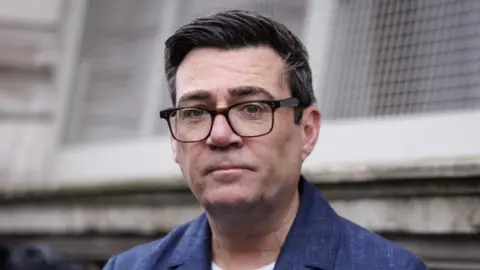 Andy Burnham has dark hair, dark glasses, blue jacket and white T-shirt