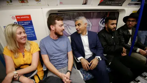 Reuters Passengers and Sadiq Khan sit together and chat inside the carriage, with Tube safety notices and advertisements visible in the background.