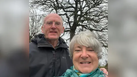 Philip George Philip George wearing a black coat and glasses in front of the large oak tree. He is stood next to his wife who has grey hair and is smiling while wearing a blue coat and scarf.