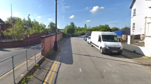 What looks like a slip road going up a hill, parting with a main road. It goes towards a train station which is out of view. Vans and cars are parked on one side of the road, with double yellow lines on the other side.