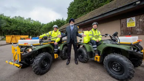 Councillor Bhupinder Gakhal with grounds maintenance team Andrew Harrison Matthew Sheffield. Mr Gakhal is stood in a grey suit between Mr Harrison and Mr Sheffield who are on green quad bikes. 
