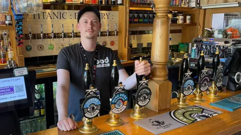 A man wearing a dark grey t-shirt and a cap stands behind a bar. There are six beer pumps on show, all with the brewery name Titanic printed on the pump clip. There are another eight beer taps on the wall behind him.