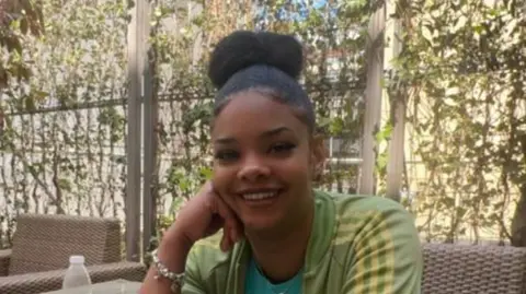 Cher Maximen is pictured smiling at the camera. She is wearing a light green Adidas top with yellow stripes down the side. Her hair is pulled up in a bun and her right hand is resting on her face, with a silver bracelet on her wrist. In the background are a green vine-type plant.