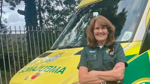 Sally Pattie, 67, has mid-length brown hair and is wearing her paramedic uniform while leaning against an ambulance. She is smiling with her arms crossed. 