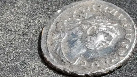 A close-up photograph shows an aged silver coin resting on a dark textured surface. The coin features a worn profile portrait surrounded by an inscription around the edge, although much of the detail has been softened over time. Visible wear and slight damage along the rim suggest the coin has been buried or handled extensively. The image highlights the coin's historical character and archaeological significance.