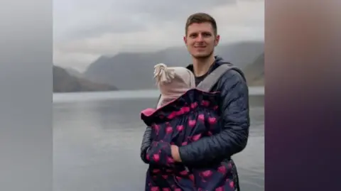 Jacob Nicholl stands in front of a lake with a mountain shrouded in mist in the background. He has short dark hair and is wearing a dark blue puffer jacket. He is holding a young child in a dark purple and pink pouch in front of him, with the child - whose face cannot be seen - wearing a pink knitted hat.