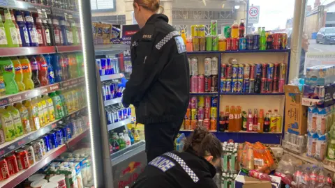 Two female police officers are examining shelves of canned and bottled drinks. One of them is crouched on the floor looking at something.