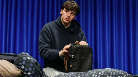 A young person sits on a bed with patterned bedding, wearing a dark hoodie and light trousers, opening a black backpack on their lap. Behind them hangs a bright blue curtain, giving the scene a staged or theatrical atmosphere.