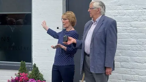 Chelsey Ward/BBC A woman in a blue top and trousers wearing a microphone headset making a speech, standing next to a man in a grey blazer and white shirt. They are standing in front of a white building.