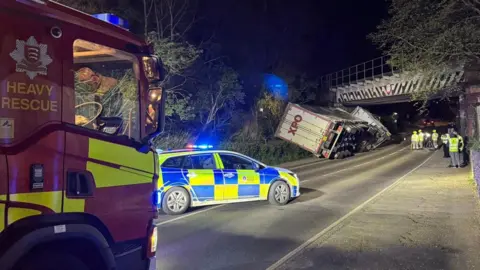 A fire truck, police car and white lorry are visible on the road at night-time. The lorry is leaning at a 45-degree angle at the entrance to the bridge.