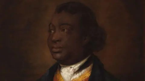 National Gallery of Canada Thomas Gainsborough, Ignatius Sancho, 1768. It is a close-up of his head and shoulders. He is looking towards his right and has his hair pulled back in a short ponytail. He is wearing a dark jacket over a yellow waistcoat with a white neck cloth tucked into it.