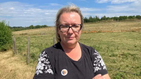Photograph of Amanda Coleman from the Chair of the Gibfield Preservation Group. She is pictured in front of open fields, with trees in the background.
