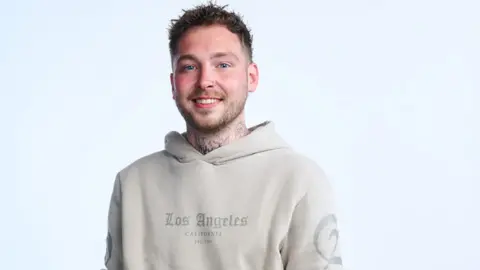 Ben is wearing a beige hooded top and has tattoos on his neck and had short brown hair.