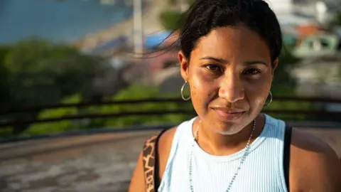 BBC A young woman with dark hair looks into the camera. She is wearing hoop earrings and a light-coloured vest top.