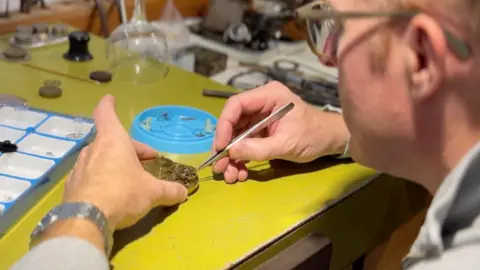 BBC Watchmaker Roger Smith is wearing glasses at a desk with a yellow covering. He's holding a partially made gold pocket watch in his left hand and using tweezers to place a mechanism in it, in his right hand.