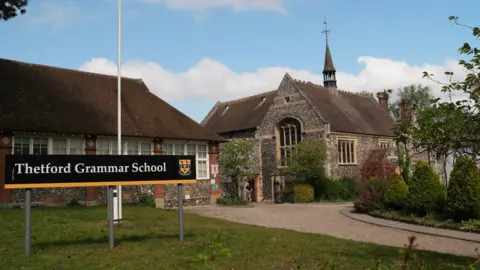 View of Thetford Grammar School. It is a flint dappled building with a spire like a church. Next to it is another long brick building with white windows. 