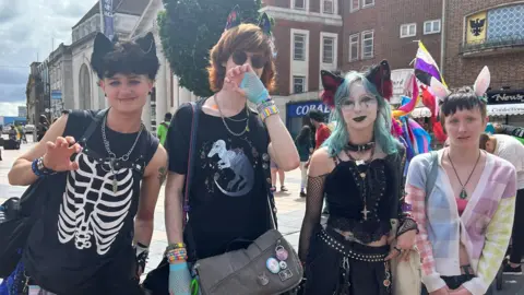 A group of four young people dressed in black, all wearing cat ears, pose in a city square. Civic buildings and shops can be seen behind them. It is a cloudy day, but bright.