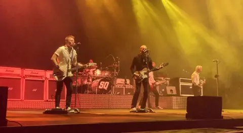 Images shows band mates singing into microphones and playing guitars. The stage has red and yellow lights shining on it