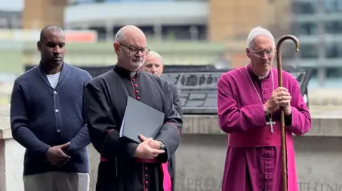 Living Bankside A number of people stand on a the walkway. One wears a bright pink ceremonial robe and holds a staff, while others in dark formal outfits stand nearby in a moment of reflection. 