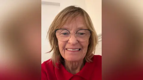 A woman wearing glasses with round frames smiling at the camera. She has blonde hair and is wearing a red collared jumper.