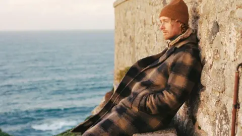 Alex Hill is wearing a brown beanie hat and a long brown and black chequered coat. He is sitting on a bench beside a sandy brick wall, looking out at the sea, which is visible behind him. The light is soft and slightly orange, appearing to be dusk.
