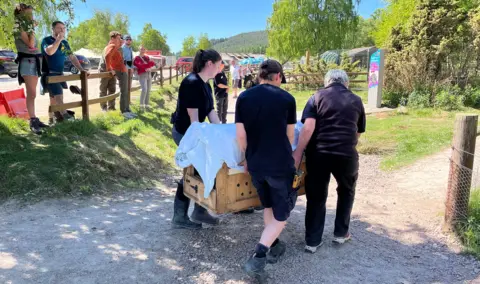 Three people in black outfits carry a wooden crate with air holes in the side. A Cloth is draped over the top and onlookers are watching
