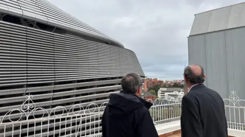 Two men stand with their back to the camera from the roof of a building looking at the stadium in the front directly