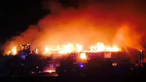 Karen Avens A large building with its roof engulfed in flames at night.