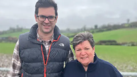 Timothy Smith and Louise Skelly. Timothy has dark hair, wearing glasses and a black water proof zip up vest and a check shirt underneath. Louse has short dark hair with grey bits, wearing pearl drop earrings and a navy zip-up fleece. Fields are in the background.