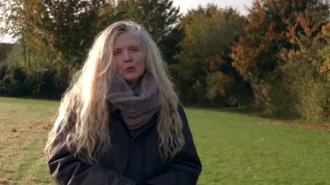 Cathy Scott, wearing a gray coat and light gray scarf, stands in a field with trees in the background. She has long hair and nose piercings on both nostrils.