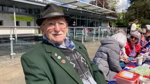 Sally Fairfax/ BBC An elderly man in a green blazer, patterned jumper and checked shirt smiling at the camera during a VE Day 80th celebration outside East Park Library. He is wearing a brown hat and glasses. He has a grey moustache and his blazer had three pin badges on. Behind him, people are sat at a long table covered in Union Jack bunting and refreshments.