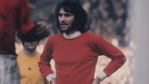 BBC Dark-haired George Best, with a thick black beard, has his hands on his hips while wearing a red Manchester United shirt with white collar and cuffs