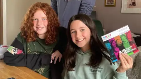 Astrid Knox has red curly hair and is wearing a a black and green hoody. Rhea Flood has long dark hair and is wearing a sage green hoody. They are sitting at a kitchen table holding a children's commissioner report entitled 12 rights for children. 